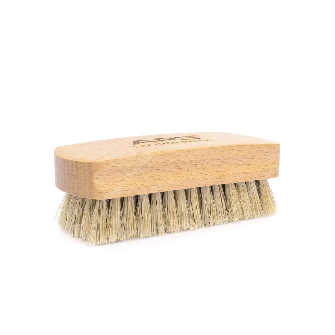 A small rectangular leather and alcantara brush with a light natural wood handle and beige bristles, shown on a plain white background. The handle features a debossed logo