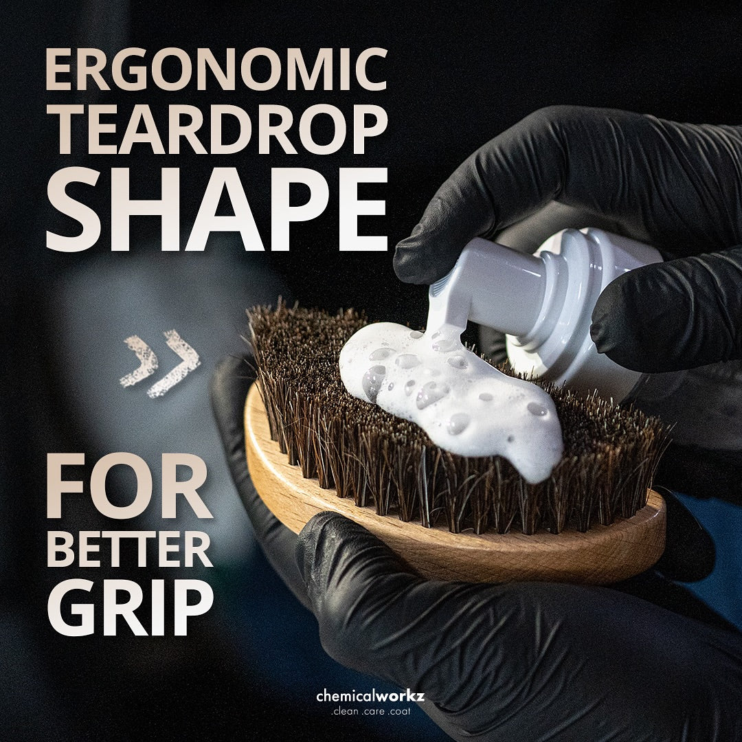 Hand holding a brush with foam and a bottle, wearing black gloves, against a dark background.