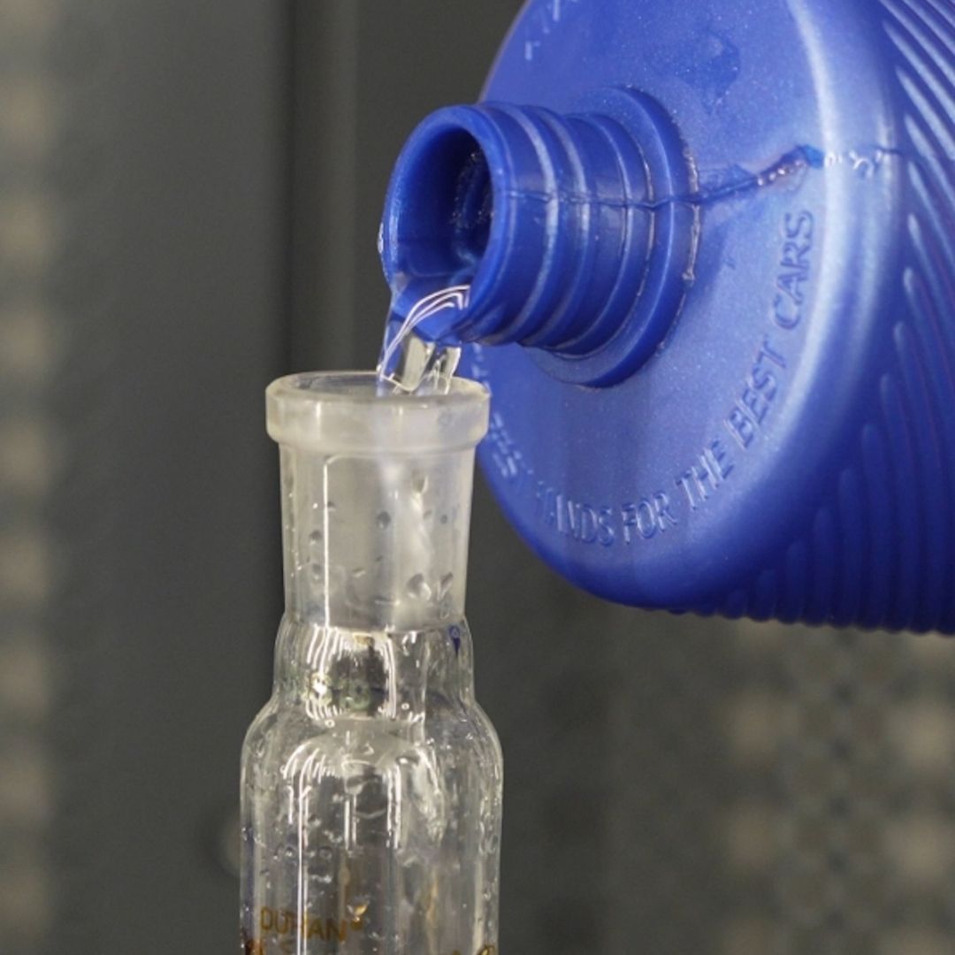A close-up of a blue plastic bottle pouring clear liquid into a small clear glass bottle.