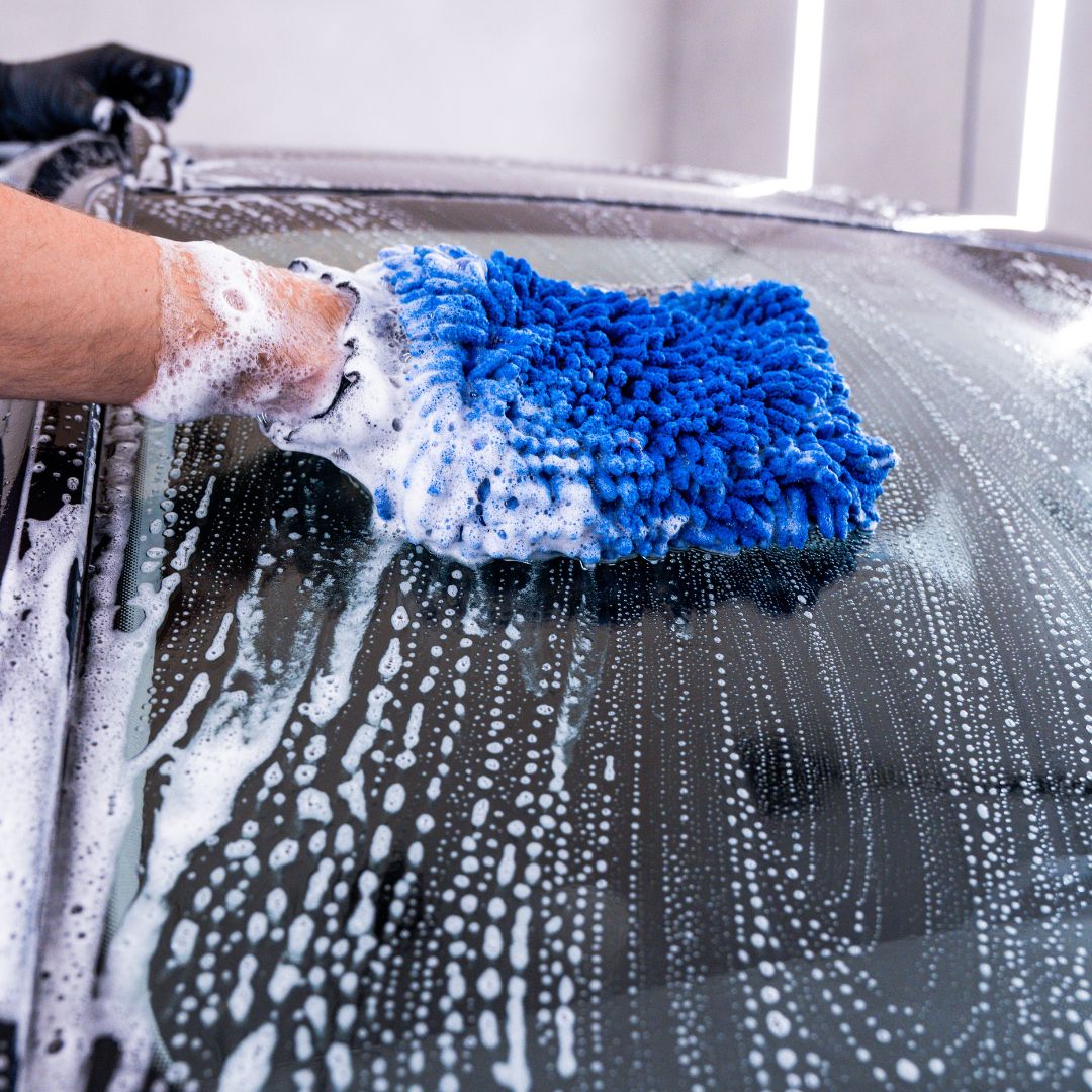 Person cleaning a car window with a blue microfiber cloth and soapy hands.