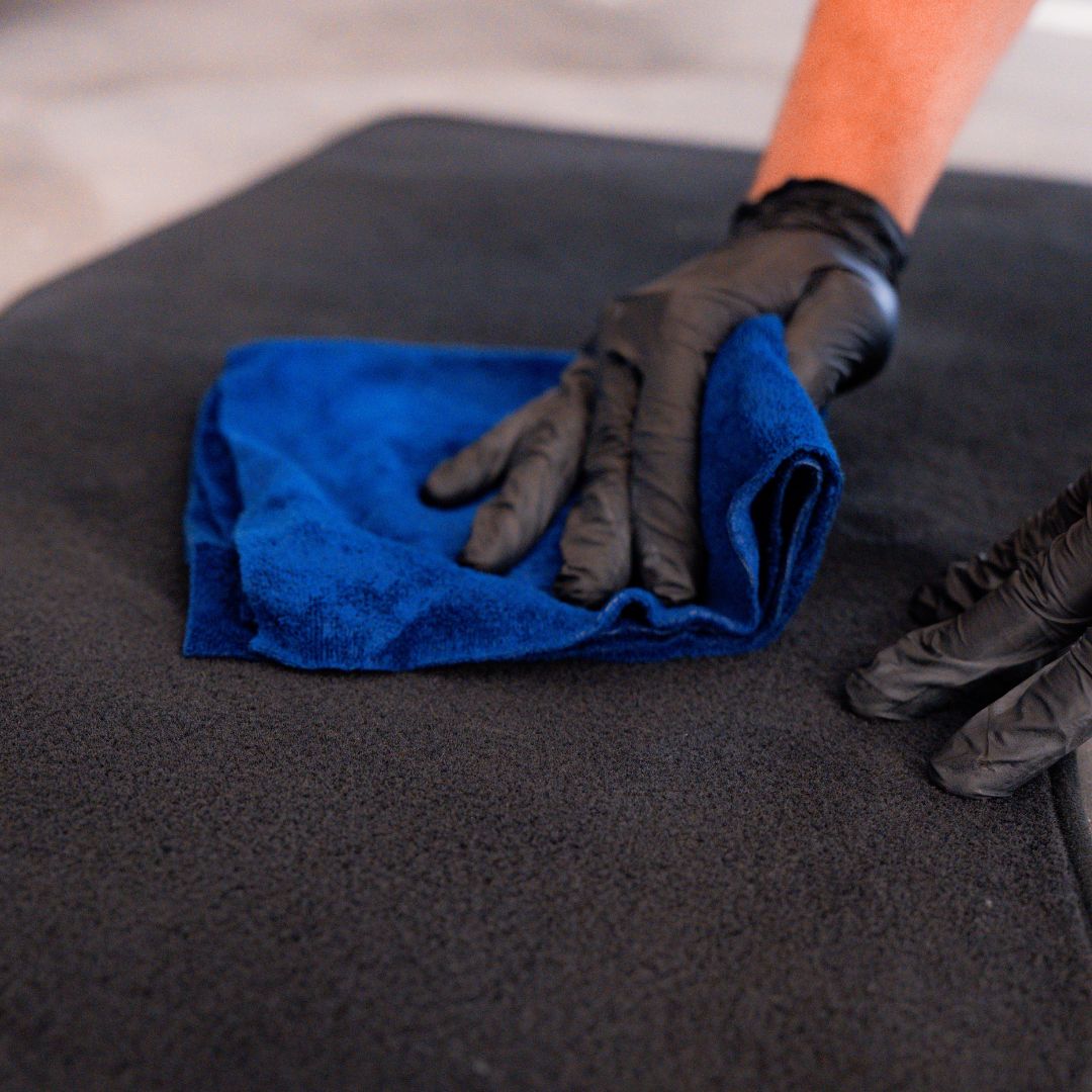 Person cleaning a car mat with a blue cloth and black gloves
