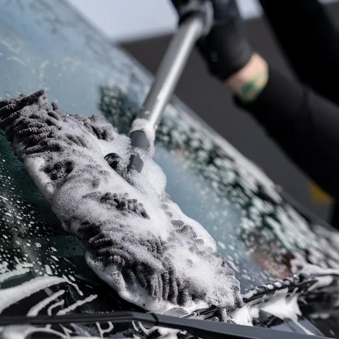 Person cleaning a car windshield with a sponge and water, covered in soap suds.
