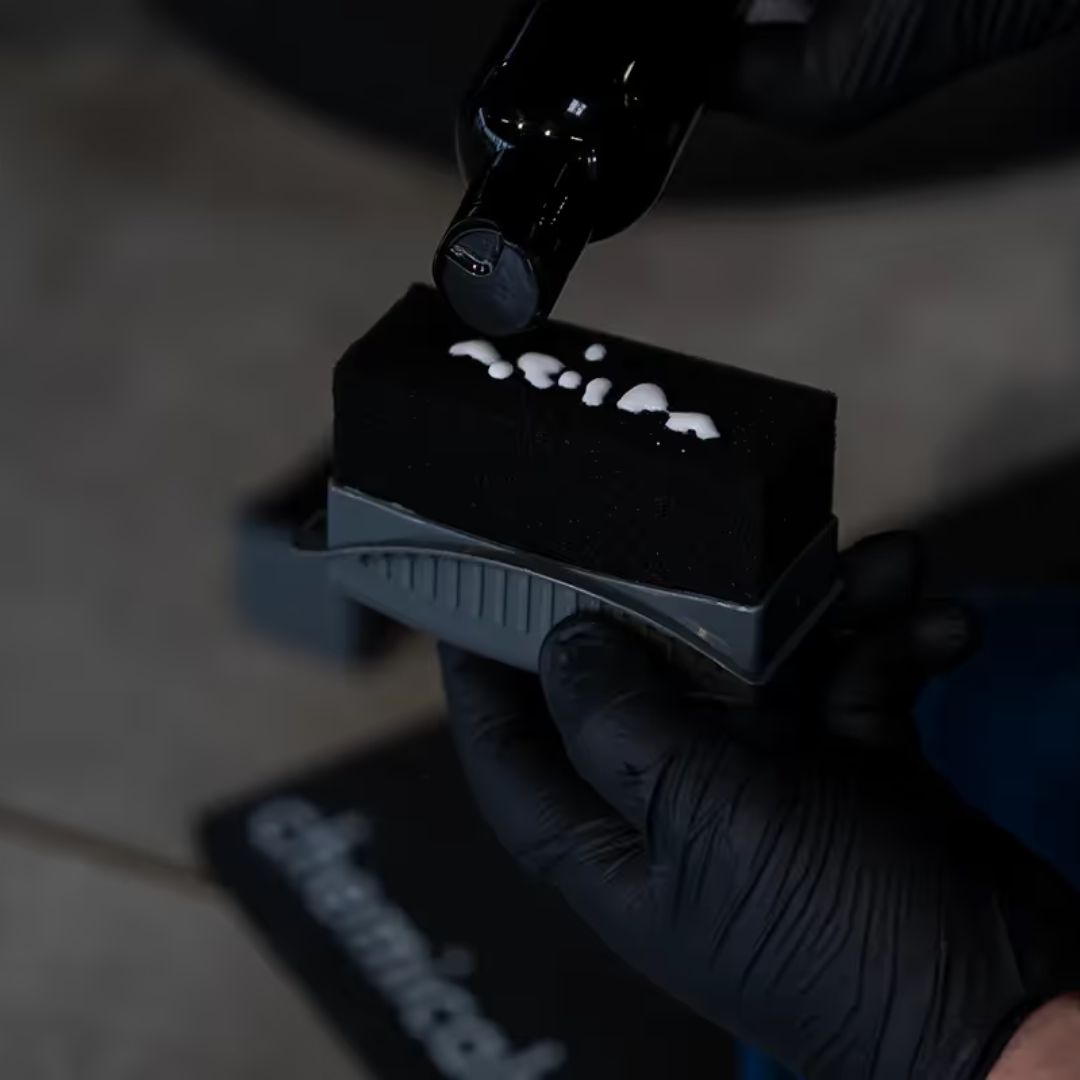 Person wearing black gloves holding a black tyre dressing applicator with white substance on a dark background