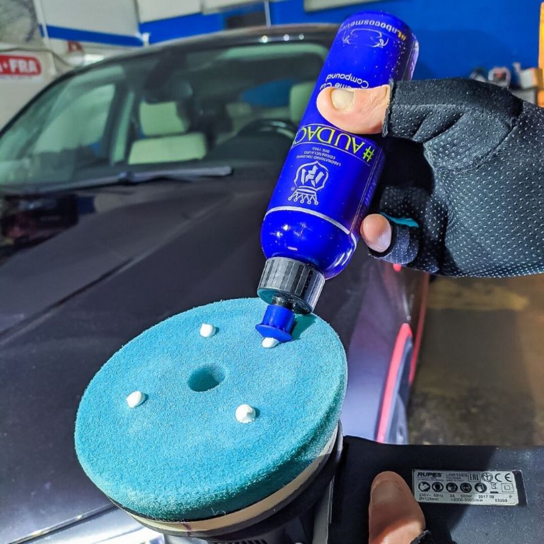 Person applying a blue bottle of Labocosmetica heavy cut compound car polish to a blue polishing pad on a car.
