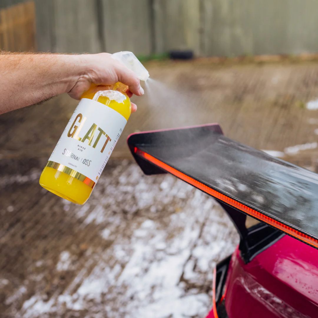 Person using a spray bottle labeled 'Stjarnagloss Glatt' on a car's spoiler outdoors.