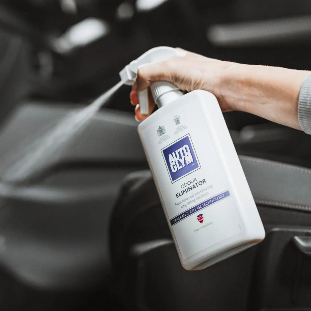 Person holding an Autoglym Odour Eliminator spray bottle inside a vehicle