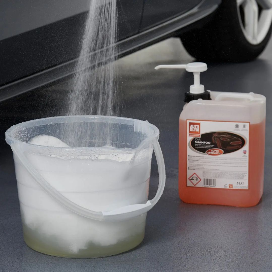 Bucket filled with soapy water and a bottle of shampoo on a car surface.