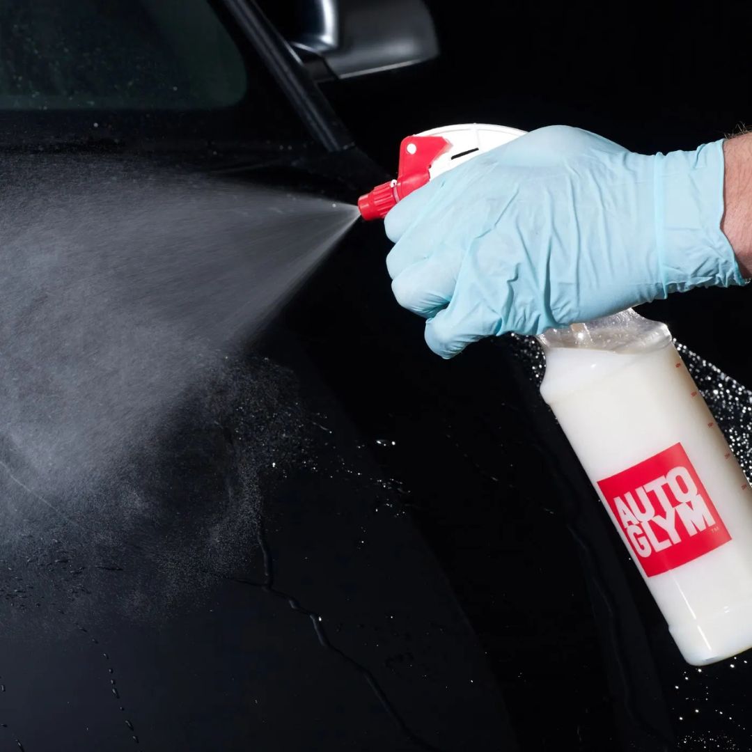 Person wearing blue gloves using a spray bottle labeled 'Glyde' on a car surface.