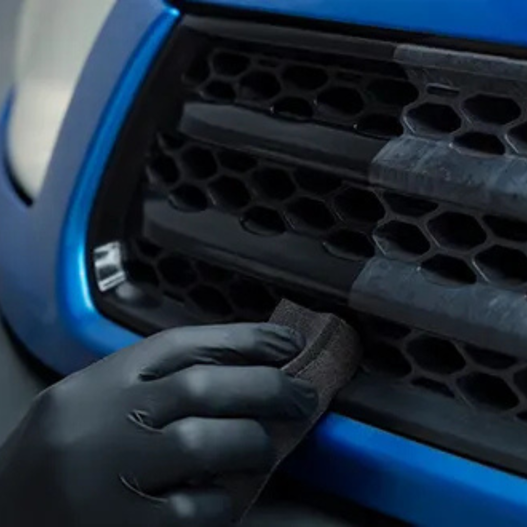 Person wearing a black glove applying a trim restorer to a car's grille.