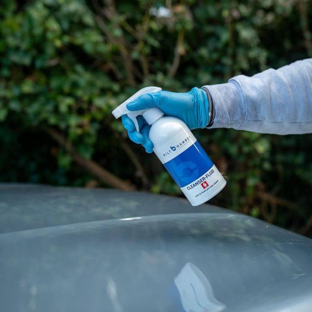 Person wearing gloves holding a spray bottle labeled 'Bilt Hamber Cleanser Fluid' over a car surface.