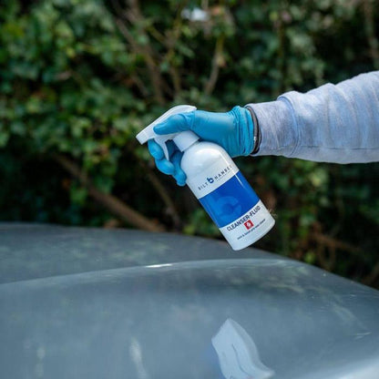 Person wearing gloves holding a spray bottle labeled 'Bilt Hamber Cleanser Fluid' over a car surface.