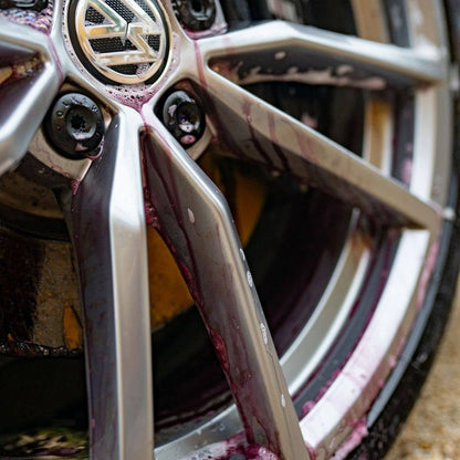 A close-up of a silver multi-spoke alloy car wheel with a metallic center cap, covered in dark purple Bilt Hamber auto-sheel, on an outdoor surface.