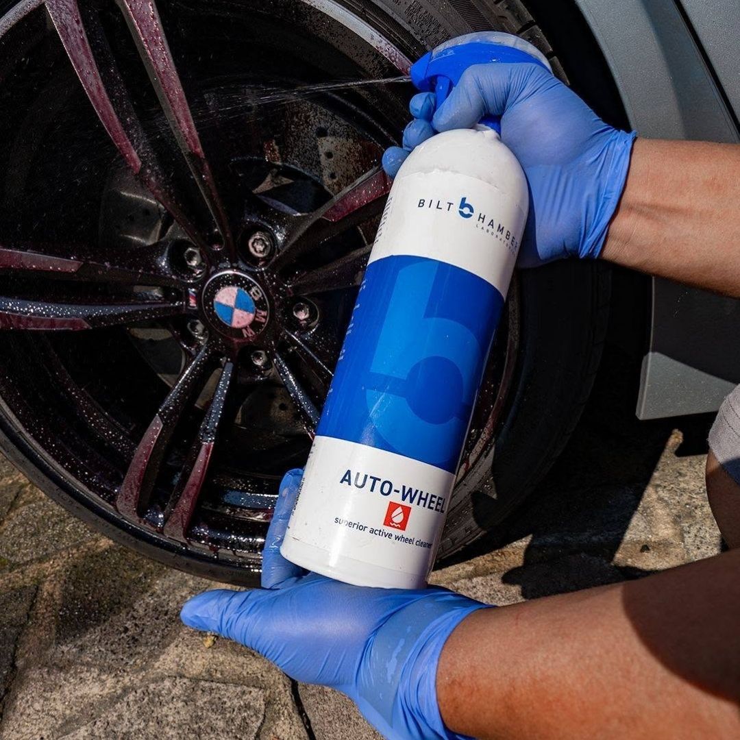 A person wearing blue gloves sprays Bilt Hamber Auto-Wheel cleaner onto a car wheel, shown in a real outdoor environment.