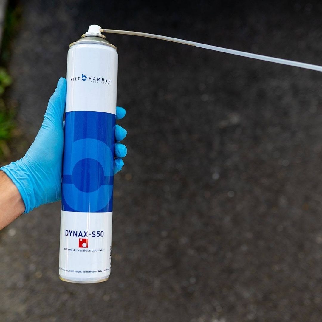 A hand wearing a blue nitrile glove holds a can of Bilt Hamber Dynax-S50 anti-corrosion paint against a dark asphalt background. The can is white with a blue circular logo and a long metal spray tube extending to the right.