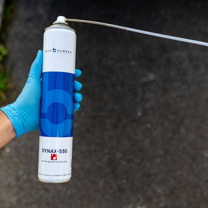 A hand wearing a blue nitrile glove holds a can of Bilt Hamber Dynax-S50 anti-corrosion paint against a dark asphalt background. The can is white with a blue circular logo and a long metal spray tube extending to the right.