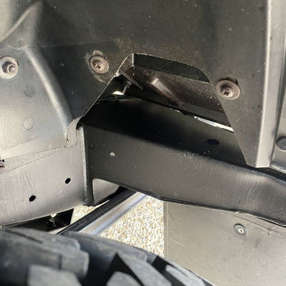 Close-up of a black metal undercarriage or frame component with visible screws and a tire edge at the bottom, photographed outdoors.