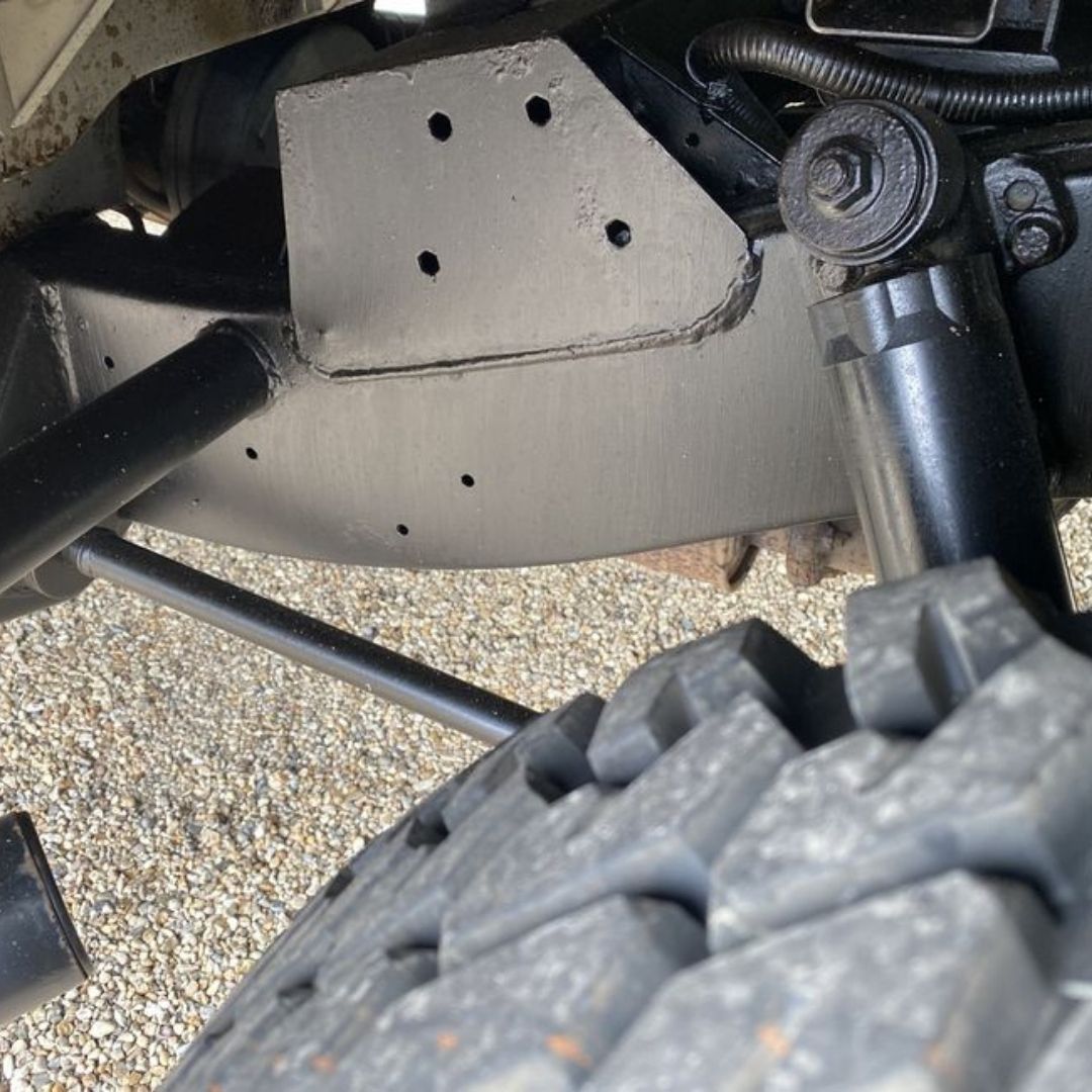 Close-up of a black metal undercarriage bracket with pre-drilled mounting holes, installed on a vehicle above a knobby off-road tire, with gravel visible in the background.