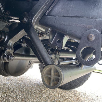 Close-up of a black metal Land Rover with Bilt Hamber Dynax UB on undercarriage, showing suspension components, exhaust, and frame parts resting on gravel outdoors.