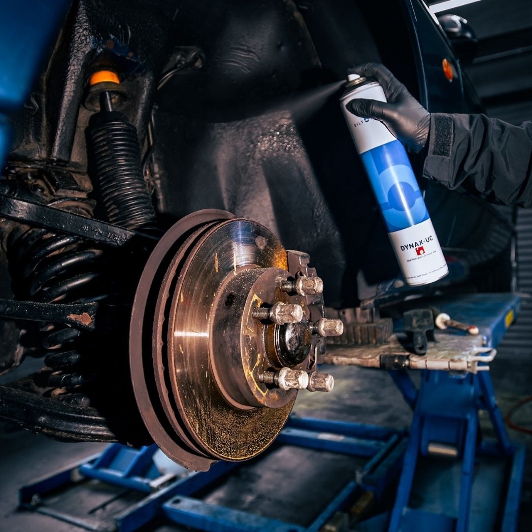 A person wearing a black glove holds a can of Bilt Hamber dynax UC rust proof in front of a vehicle's exposed brake rotor and suspension components.