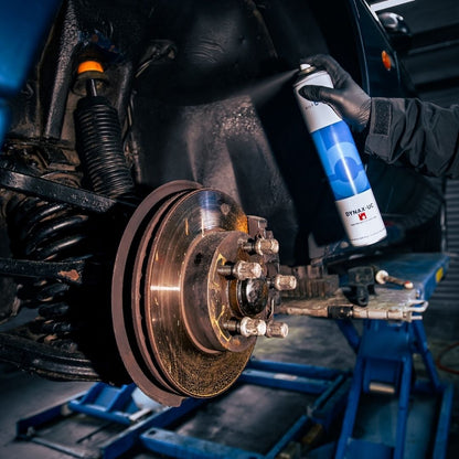 A person wearing a black glove holds a can of Bilt Hamber dynax UC rust proof in front of a vehicle's exposed brake rotor and suspension components.