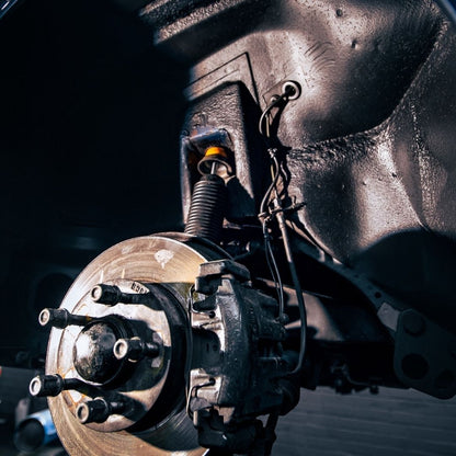 Close-up of a vehicle's wheel hub, brake rotor, suspension spring, and brake assembly against a dark background.
