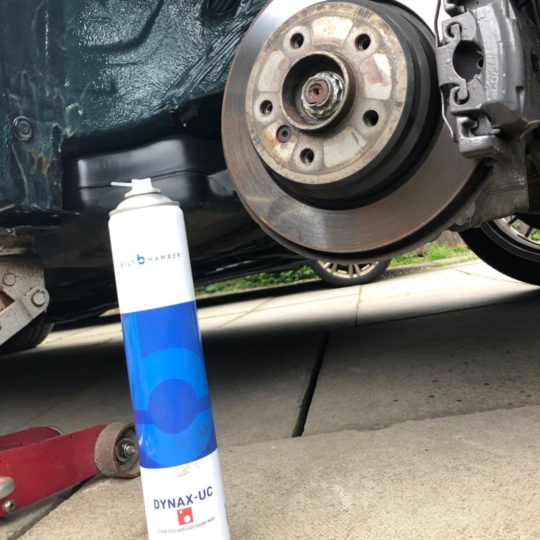 A can of Bilt Hamber Dynax-UC Rust proof is shown in a real-world garage setting next to a car wheel hub and brake rotor, indicating its use for automotive lubrication.