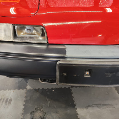 Close-up of a Volvo 480 front bumper and headlight area of a red car, showing a black license plate frame and bumper with some wear and dirt on a garage floor.