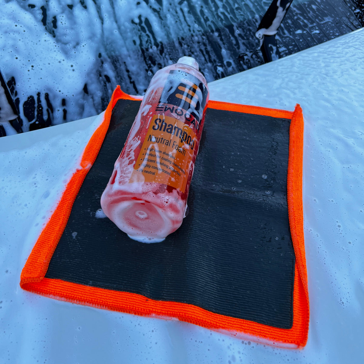 A bottle of shampoo wrapped in white foam rests on a black and orange microfiber cleaning mat placed on a snow-covered surface in front of a foamy car windshield.