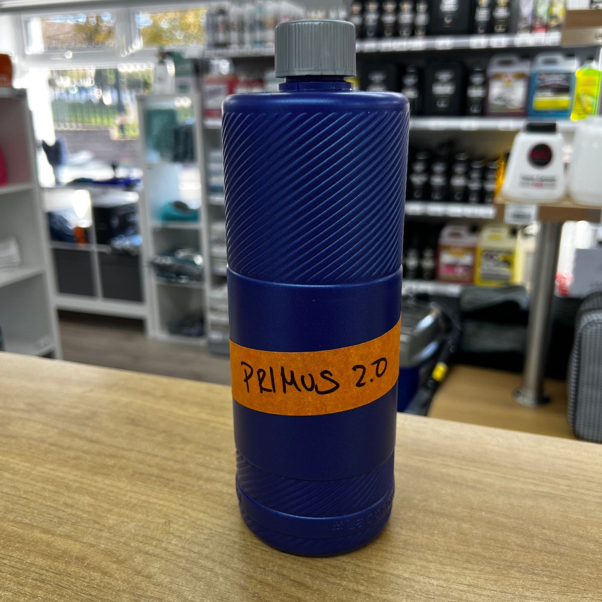 A blue cylindrical bottle with a ribbed texture and a gray cap, standing on a wooden counter in a store. An orange paper label wrapped around the middle reads 'PRIMUS 2.0' in black handwritten text.