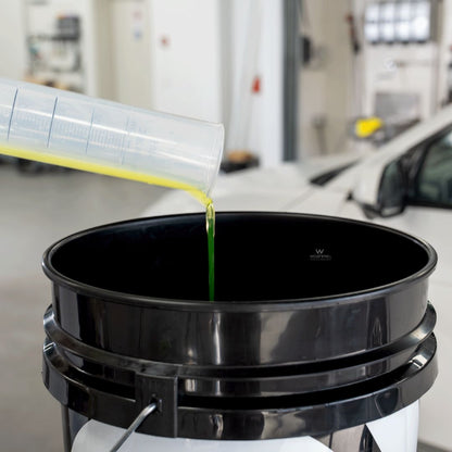 Bottle of Koch Chemie The Finisher TropicSnow being poured from a container into a black oil filter container with a car in the background.