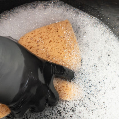 Hand wearing a black glove holding a brown sponge over a soapy water surface.