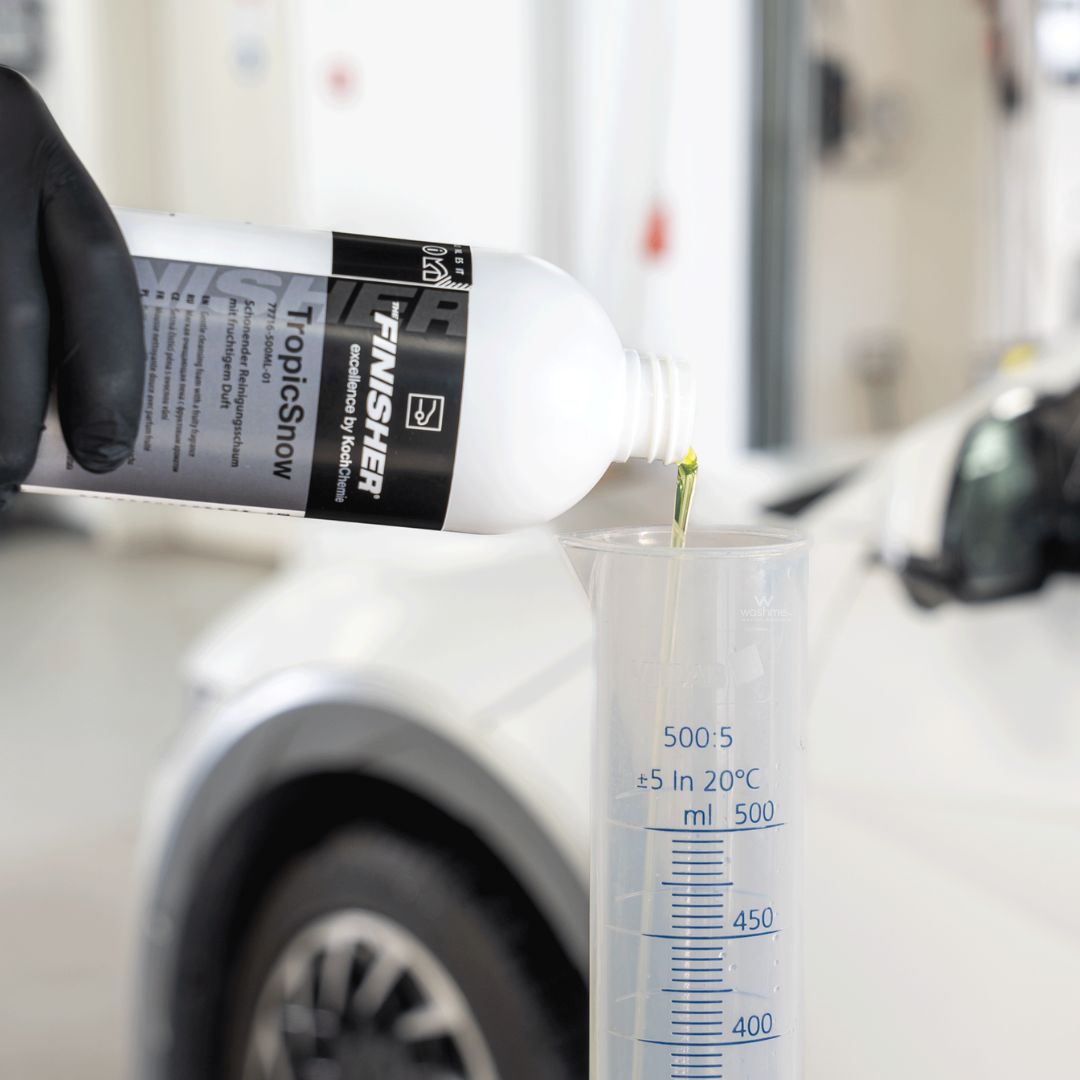 Person pouring a Bottle of Koch Chemie The Finisher TropicSnow from a bottle labeled 'FINISHER' into a measuring cup with a car in the background.