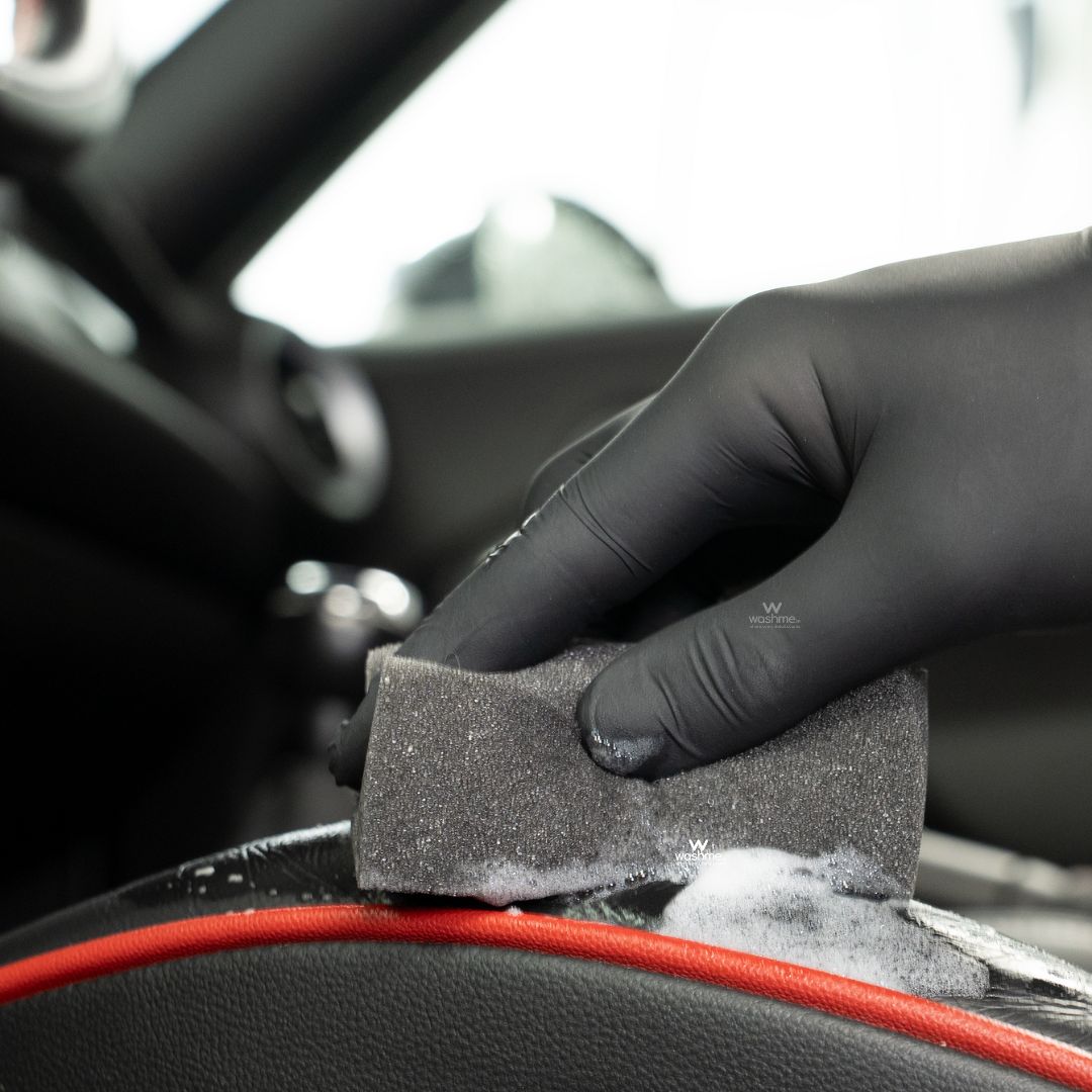 Hand wearing a black glove holding a sponge using Koch Chemie The Finisher LeatherTexClean inside a car.