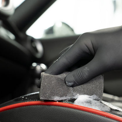 Hand wearing a black glove holding a sponge using Koch Chemie The Finisher LeatherTexClean inside a car.
