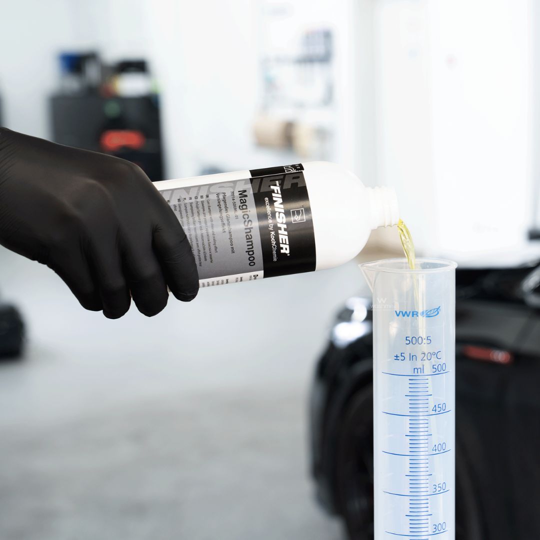 Hand in black glove pouring a Koch Chemie The Finisher TropicSnow car wash shampoo from a 'FINISHER' bottle into a measuring cylinder.