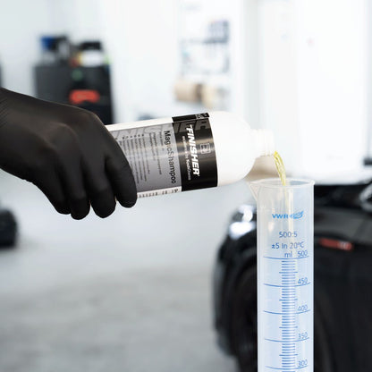 Hand in black glove pouring a Koch Chemie The Finisher TropicSnow car wash shampoo from a 'FINISHER' bottle into a measuring cylinder.