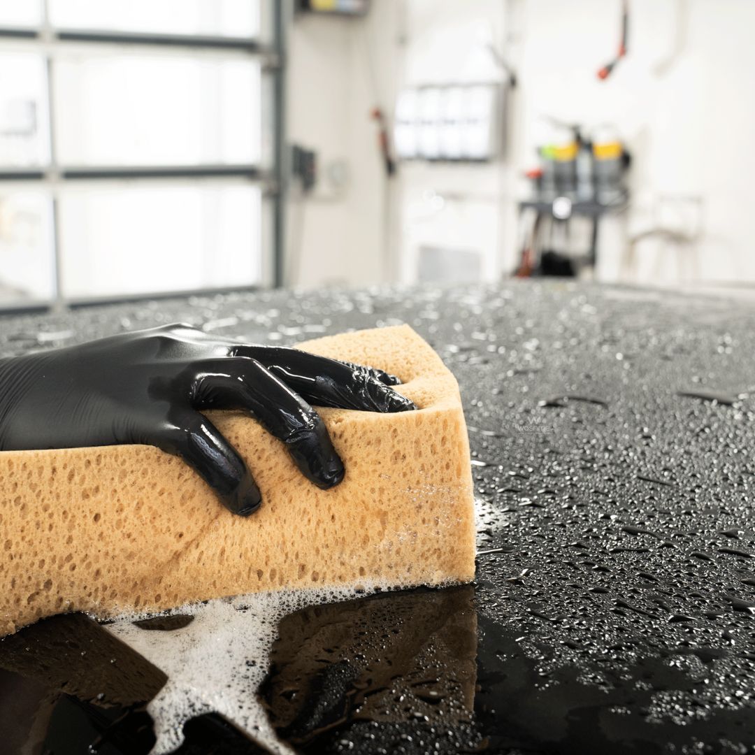 Hand wearing a black glove holding a beige sponge over a car surface with soap suds.