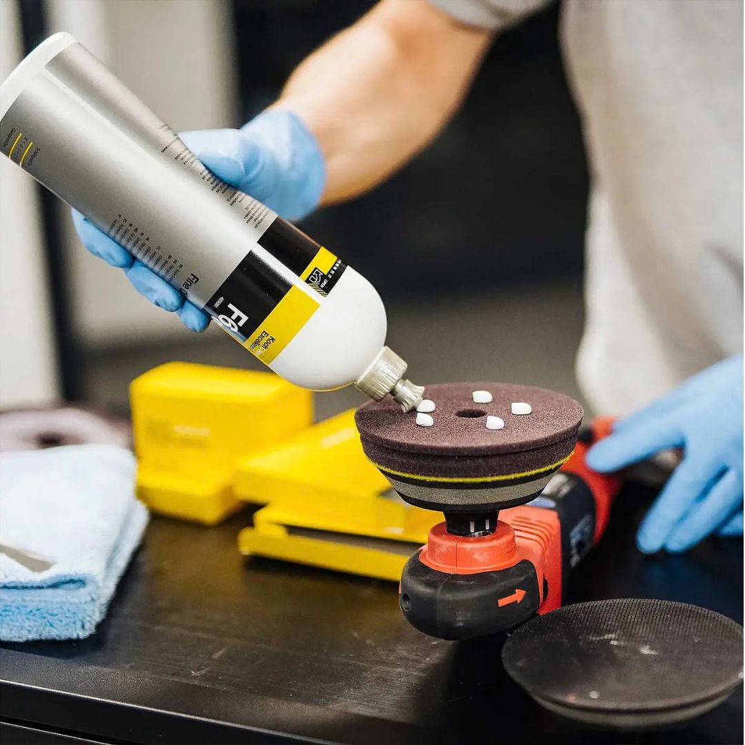 Person applying Koch Chemie Fine Cut F06.01 to a polishing pad with gloves on, on a blurred background.