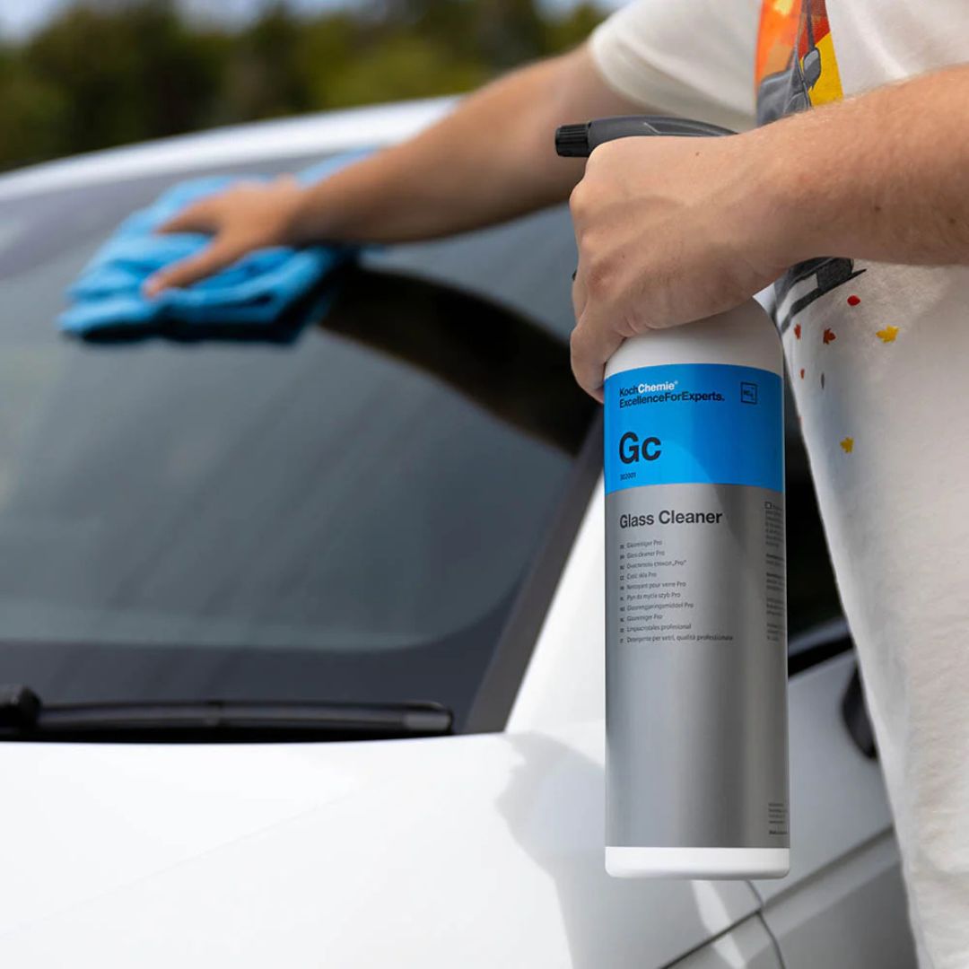 Person cleaning a car window with a spray bottle labeled 'Gc Glass Cleaner' and a blue microfiber cloth.