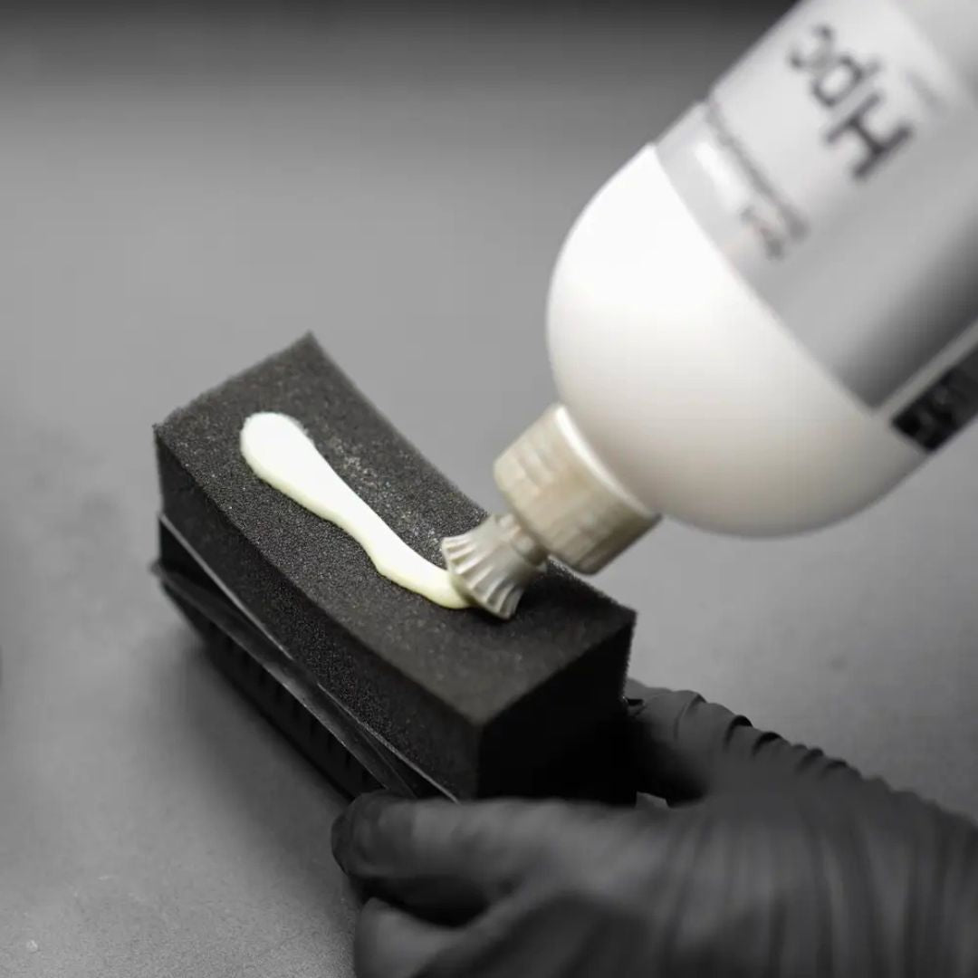 Person applying white cream to a black sponge with a bottle labeled 'Koch Chemie HPC Hydro Plast Care' on a gray background.