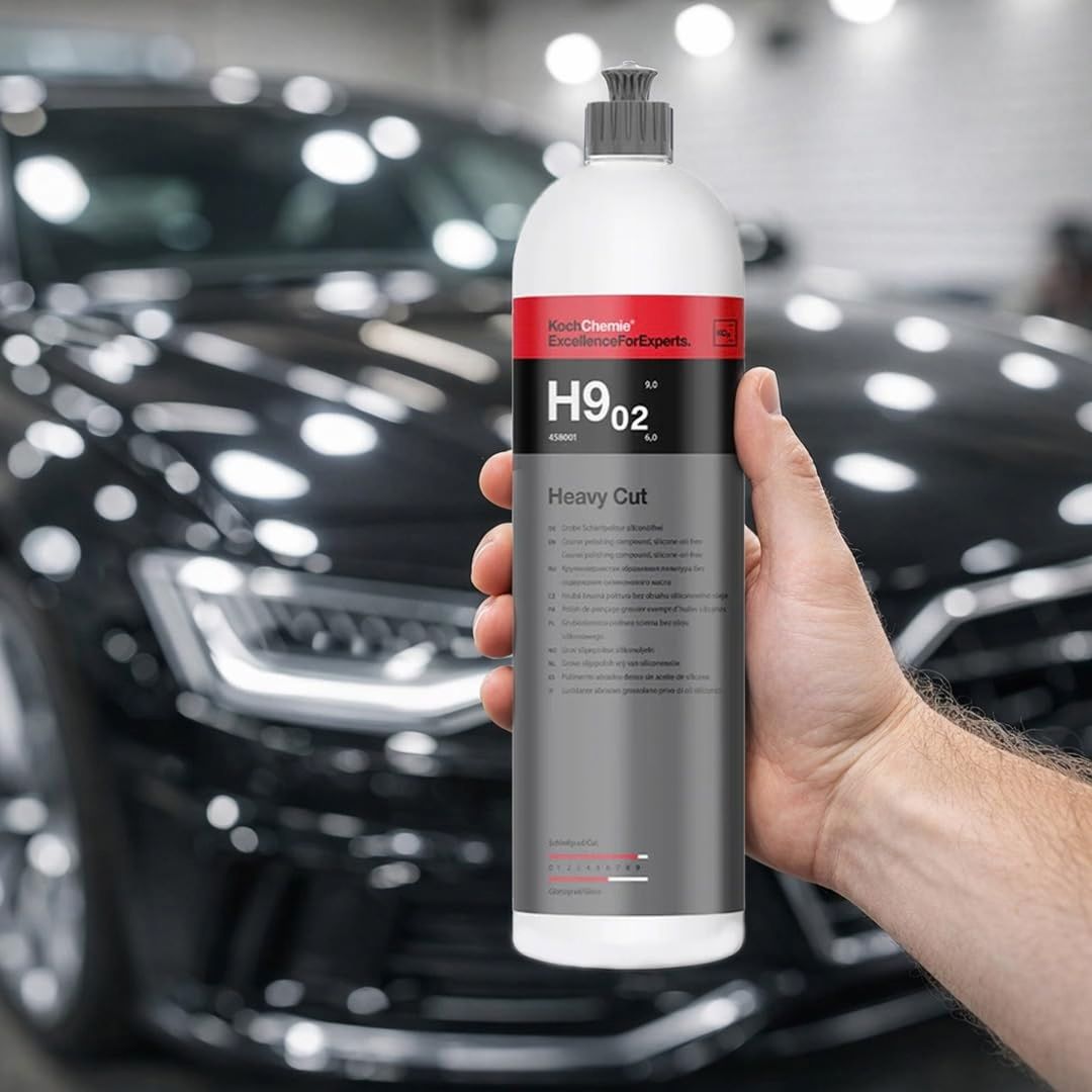 A hand holds a white bottle of Koch Chemie H9.02. Heavy Cut polishing compound against a blurred background of a shiny black car in a garage.