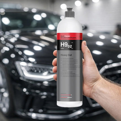 A hand holds a white bottle of Koch Chemie H9.02. Heavy Cut polishing compound against a blurred background of a shiny black car in a garage.