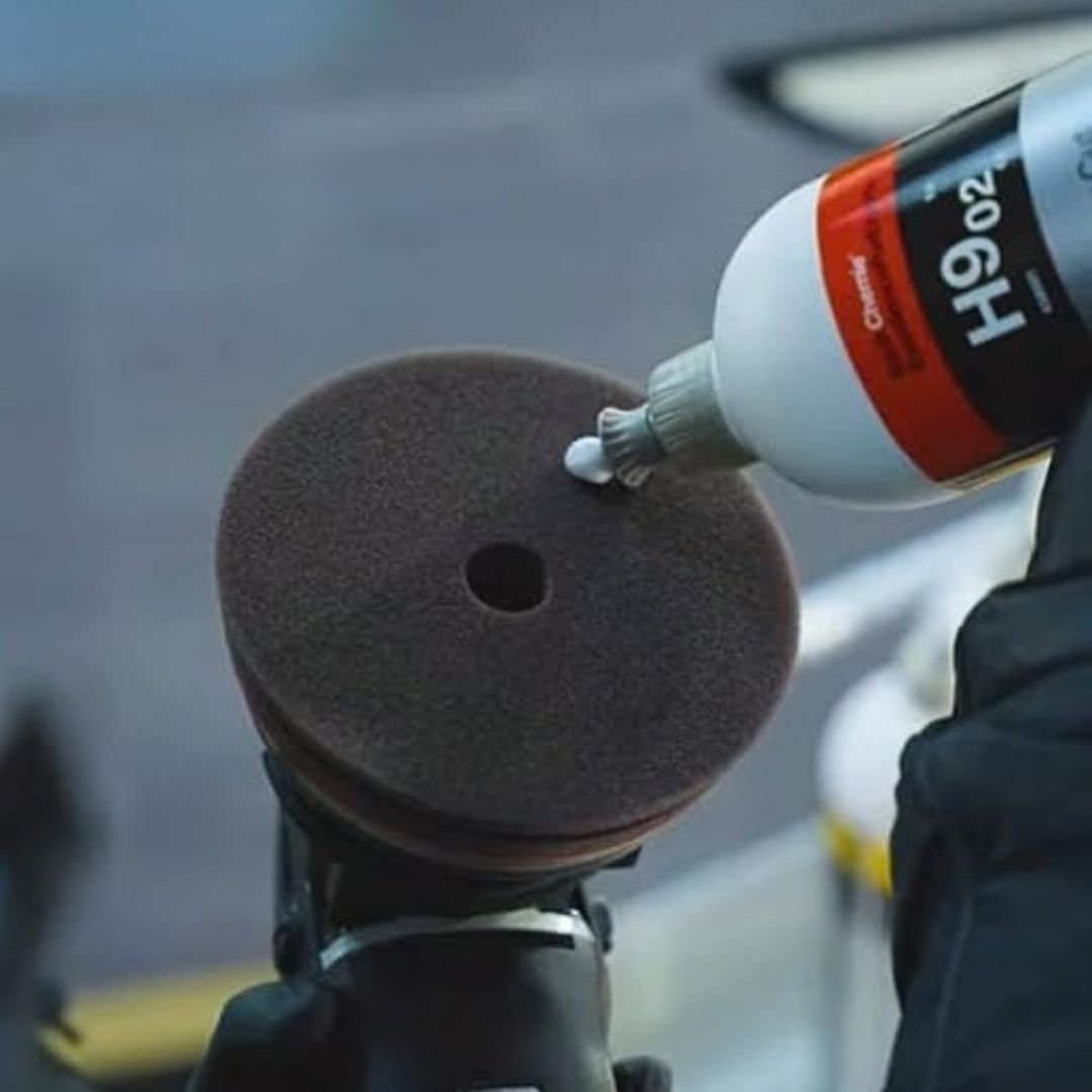 A close-up action shot of a person applying Koch Chemie “H9.02. Heavy Cut” polishing compound onto a brown foam pad mounted on a polishing machine.