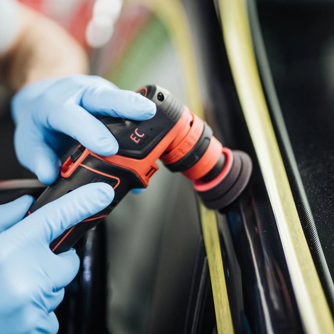 A close-up action shot of a person wearing blue work gloves holding a cordless electric polishing machine with Koch Chemie “H9.02. Heavy Cut” polishing compound