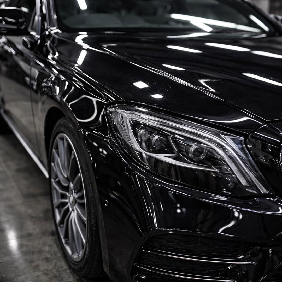Close-up of a glossy black Mercedes E-Class, showing the front headlight, hood, side body panel, and multi-spoke alloy wheel in a showroom setting.
