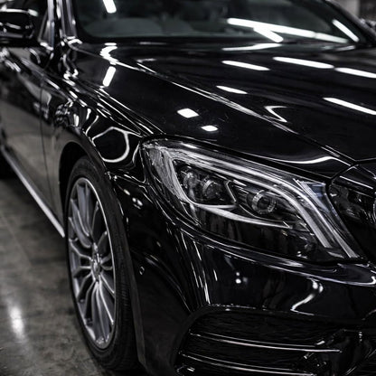 Close-up of a glossy black Mercedes E-Class, showing the front headlight, hood, side body panel, and multi-spoke alloy wheel in a showroom setting.