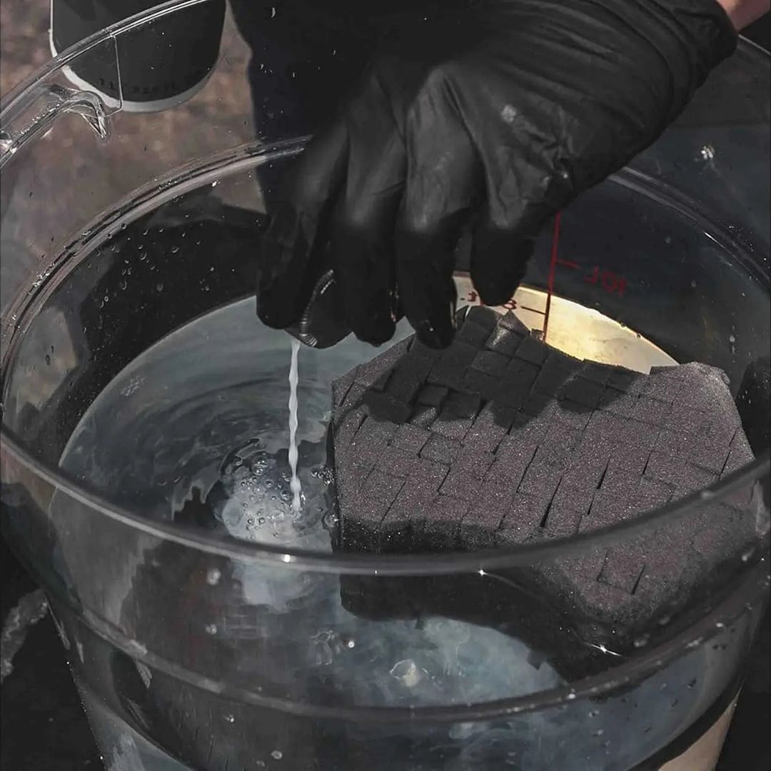 Person wearing a black glove holding a black sponge over a bucket of water.