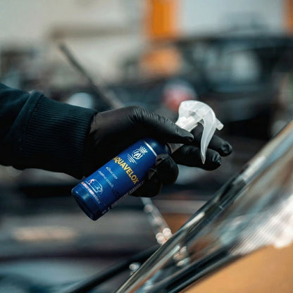 Person holding a spray bottle labeled 'Aquavelox' in front of a car window.