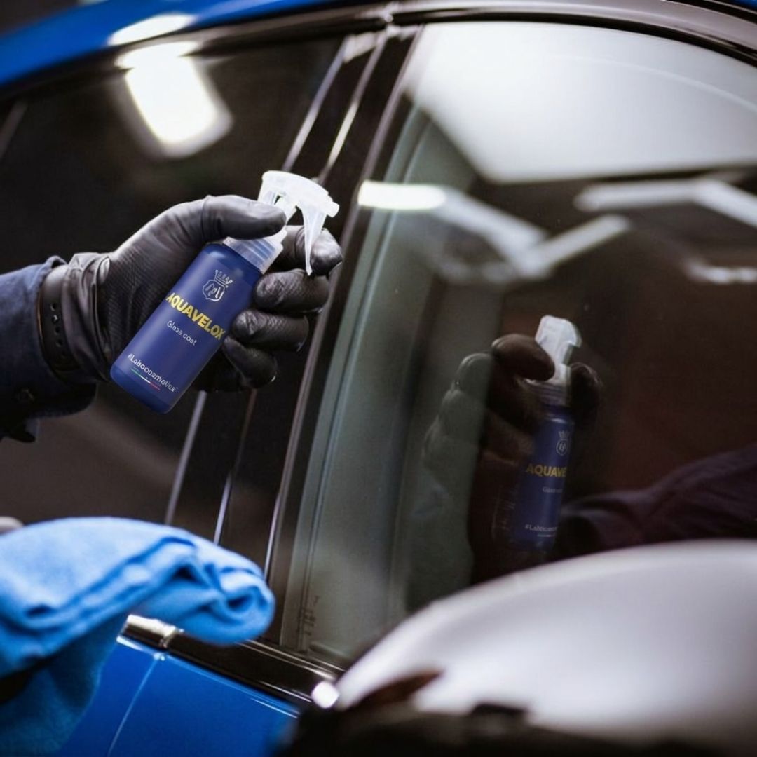 Person cleaning a car window with a spray bottle labeled 'Aquavelox' and a cloth.