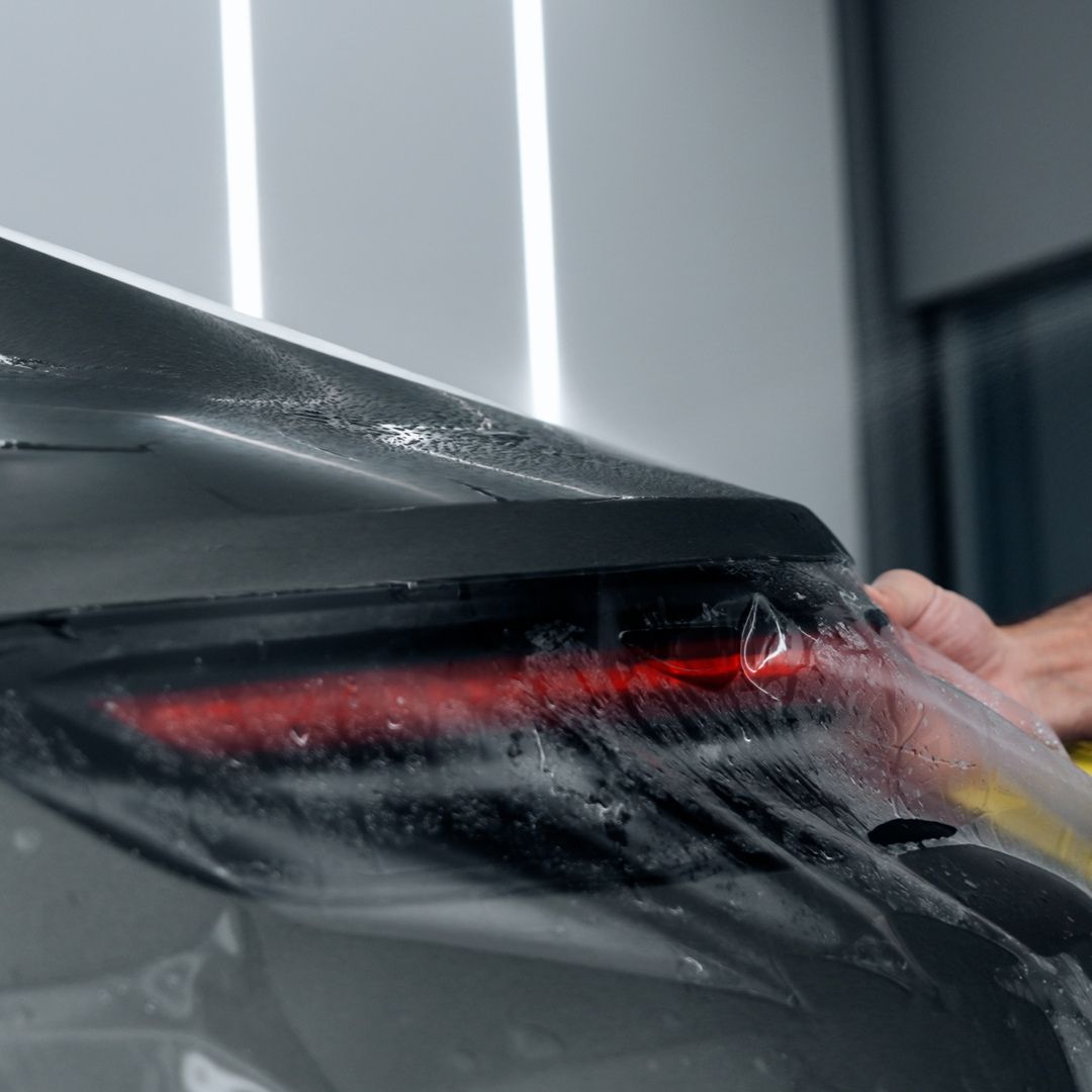 A close-up of a person applying a clear protective film or wrap to the rear quarter panel and taillight area of a dark gray car, with water droplets visible on the surface.